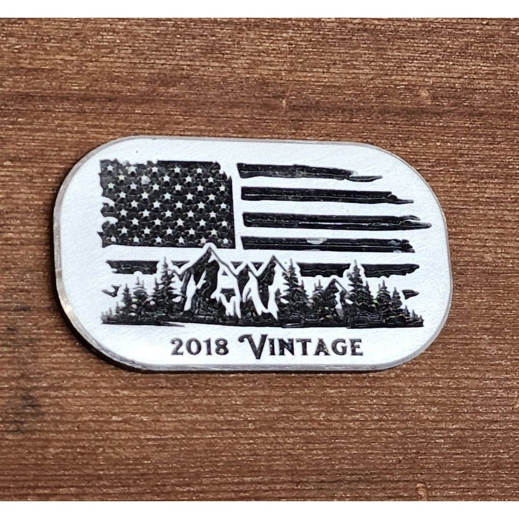 Metallic badge with American flag design, mountains, and trees on a wooden background