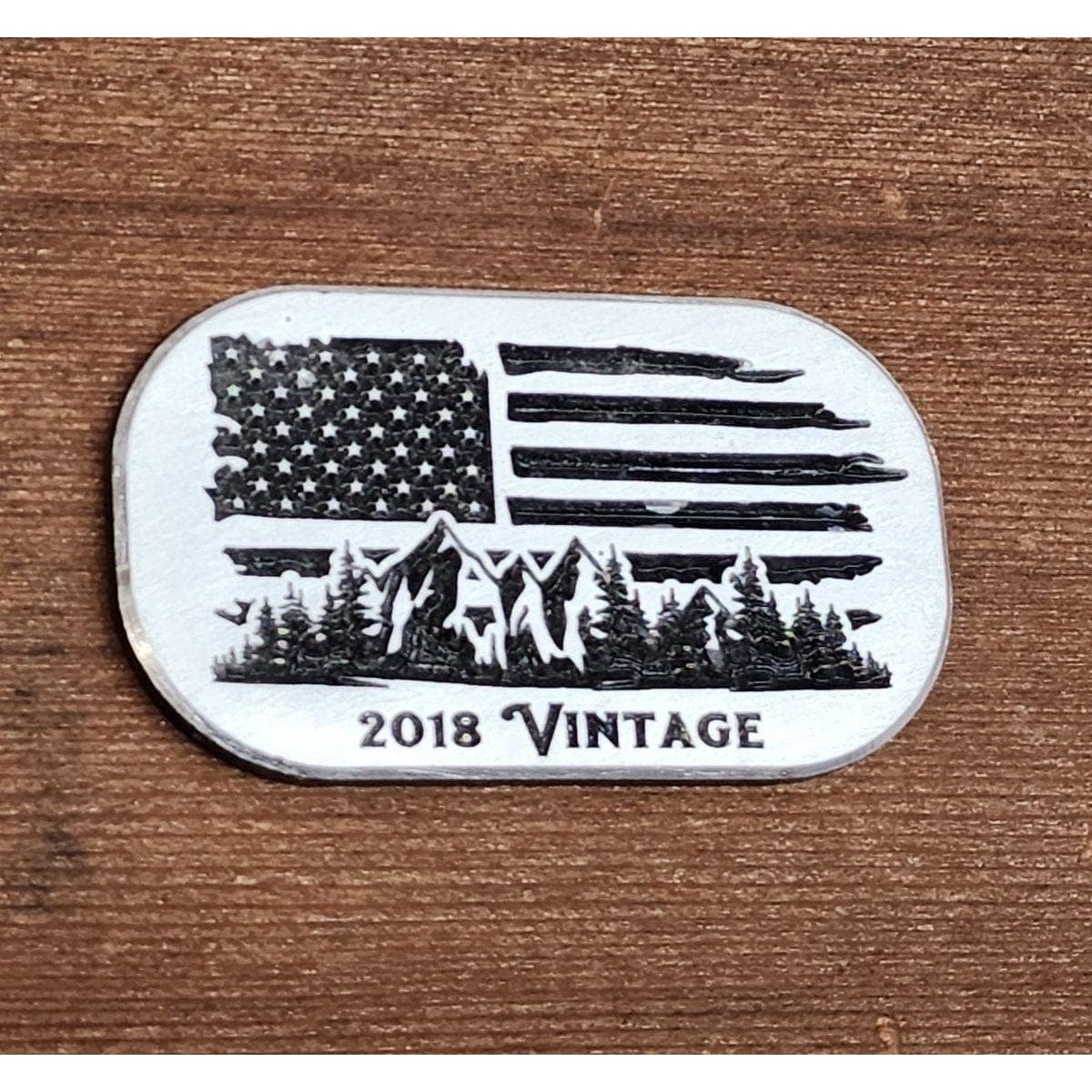 Metallic badge with American flag design, mountains, and trees on a wooden background